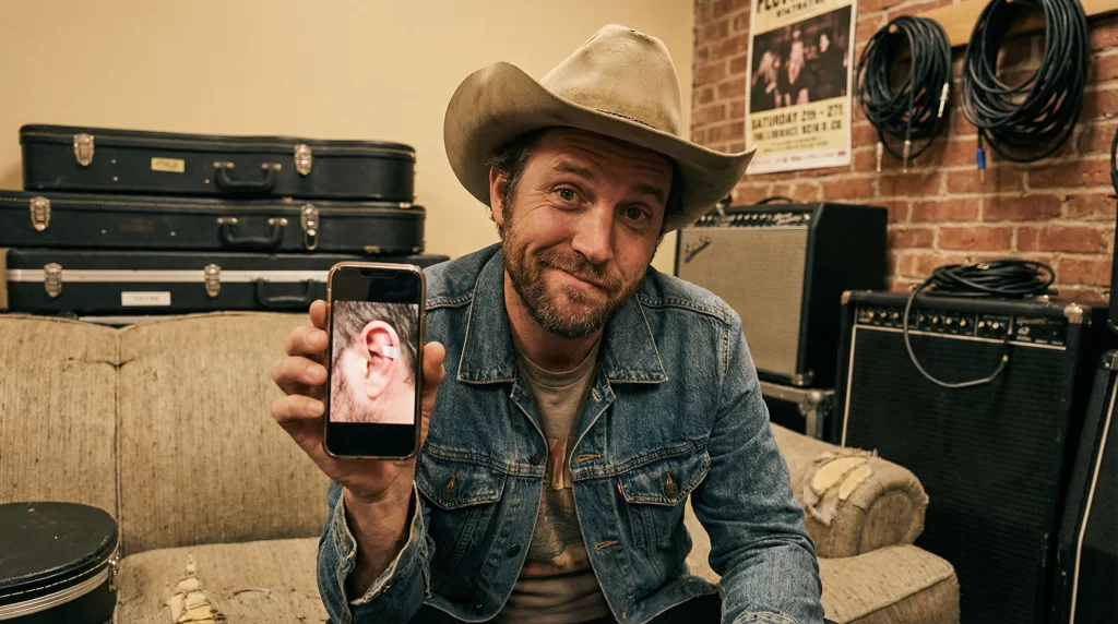 Country singer in cowboy hat sitting backstage showing phone photo of ear injury