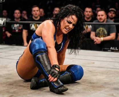 A female professional wrestler clutching her ankle in pain on the mat of a brightly lit wrestling ring