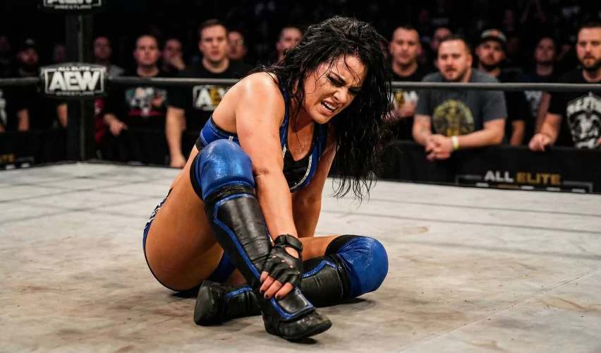 A female professional wrestler clutching her ankle in pain on the mat of a brightly lit wrestling ring