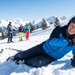 A male skier in winter gear lying on a snowy mountain slope clutching his shoulder in pain after a fall