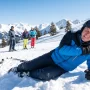 A male skier in winter gear lying on a snowy mountain slope clutching his shoulder in pain after a fall